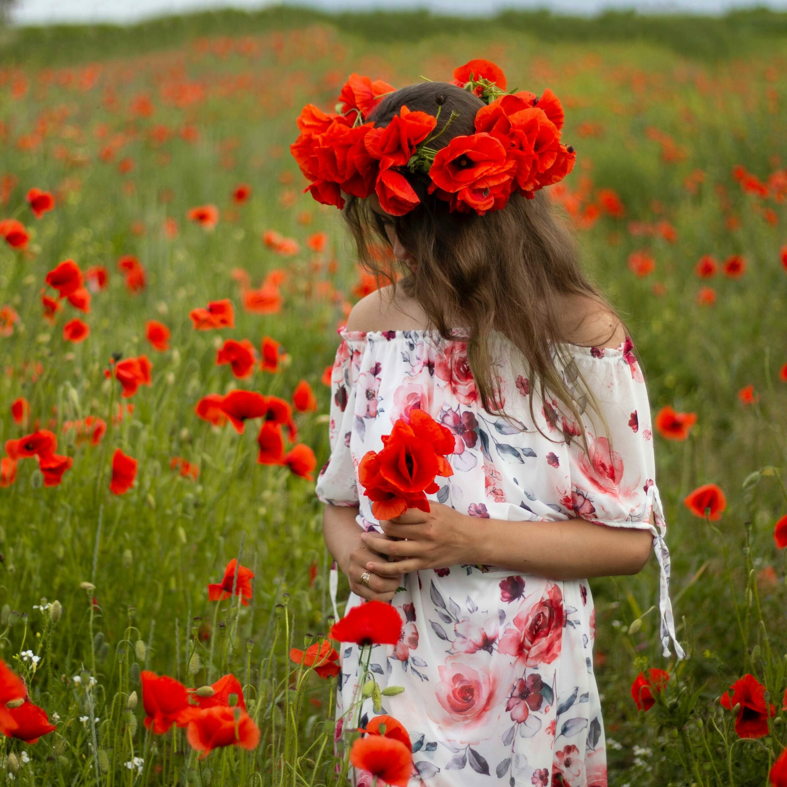 Woman standing in a field of red flowers wearing a flower crown and white floral dress.